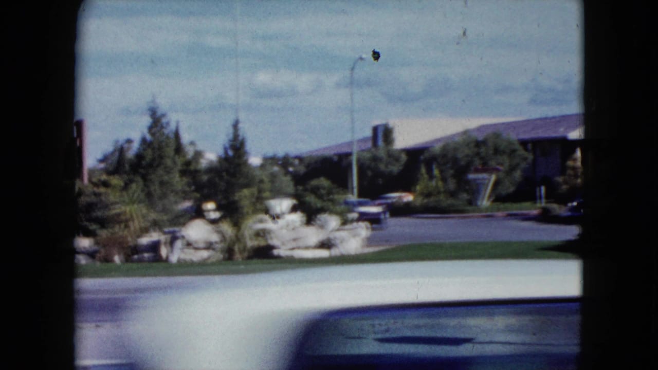 1959 Nevada street frame showing a low building, trees, rock landscaping, parked cars, roadway, and a passing car cropped in the foreground.