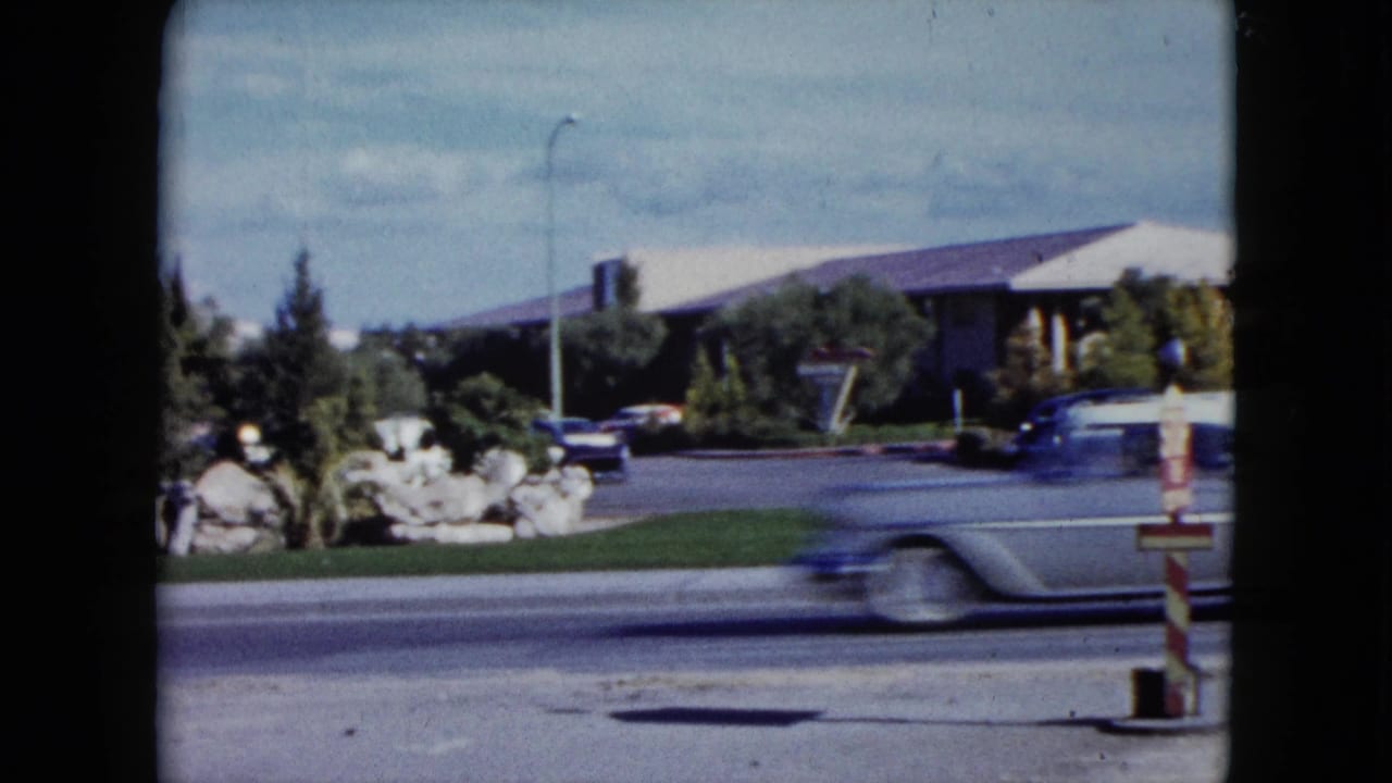 1959 Nevada home-movie frame of a car passing quickly in front of a low white-roofed roadside building and parked automobiles.