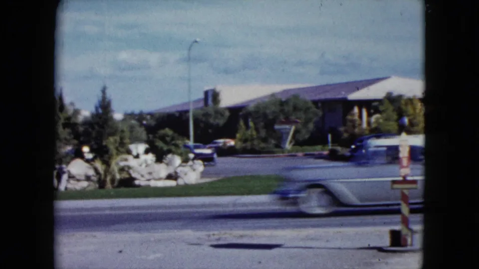 1959 Nevada home-movie frame of a car passing quickly in front of a low white-roofed roadside building and parked automobiles.