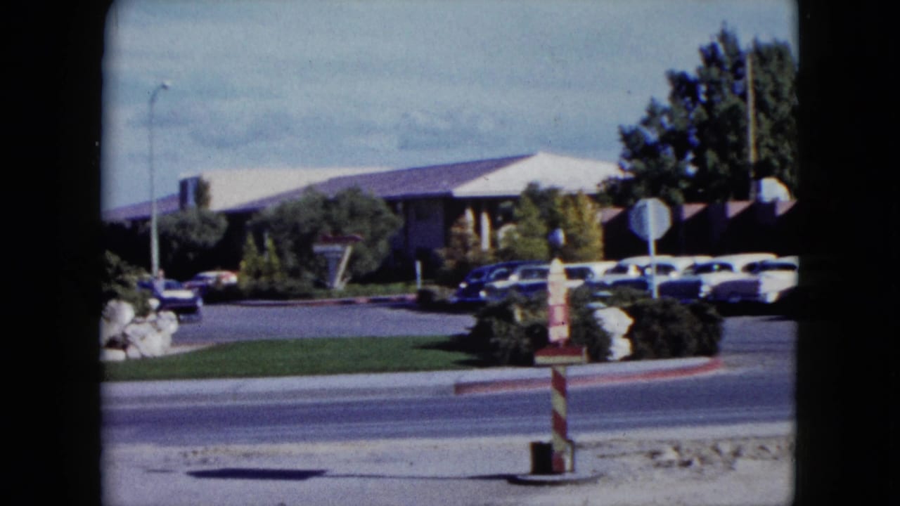 Roadside building and parking lot seen across a street with cars and a striped pole in front.