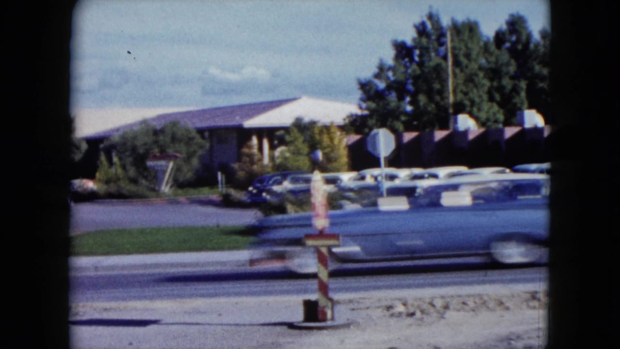1959 Nevada 8mm frame showing a large mid-century car blurred by motion in front of parked cars and a low roadside building.