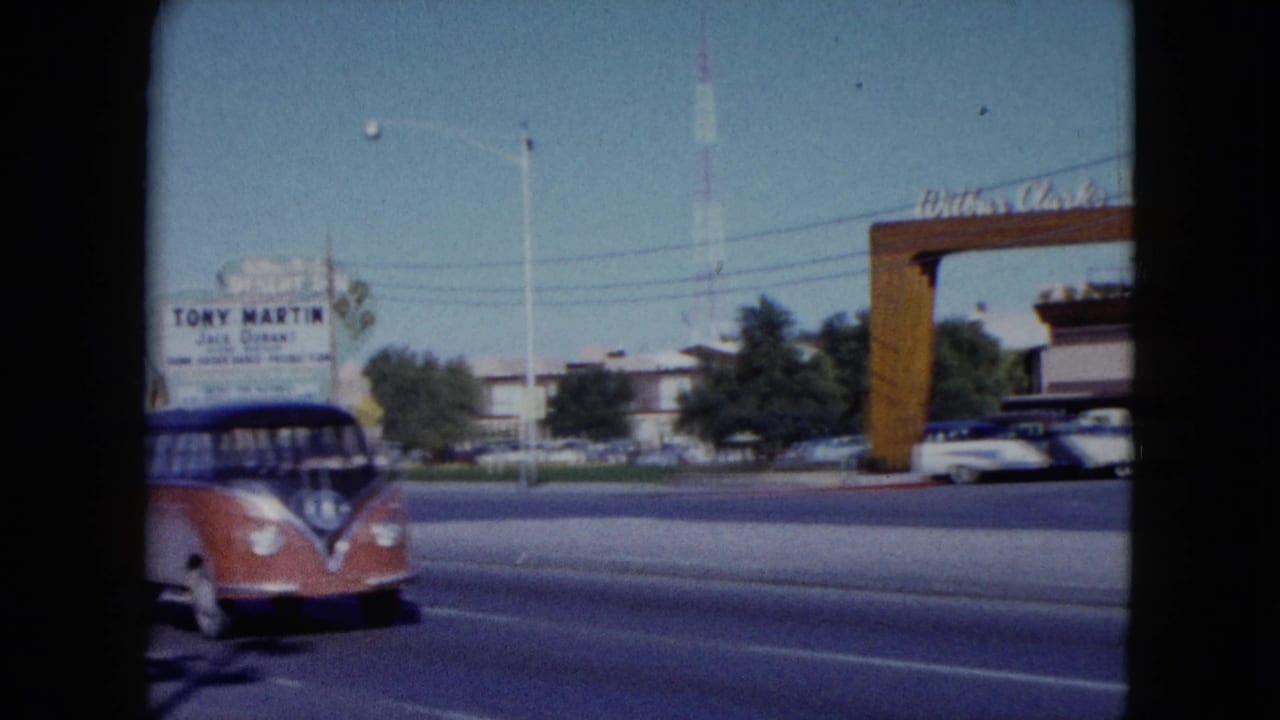A red and blue Volkswagen Type 2 bus moves along the roadway in front of the Desert Inn entrance in Nevada, with mid-century cars and a tower visible beyond.