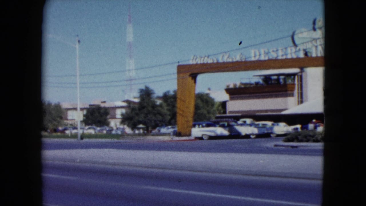 The Desert Inn entrance arch and signage stand across a sunlit Nevada roadway as 1950s cars pass and others sit in the parking area.
