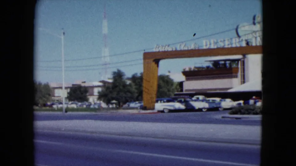 The Desert Inn entrance arch and signage stand across a sunlit Nevada roadway as 1950s cars pass and others sit in the parking area.