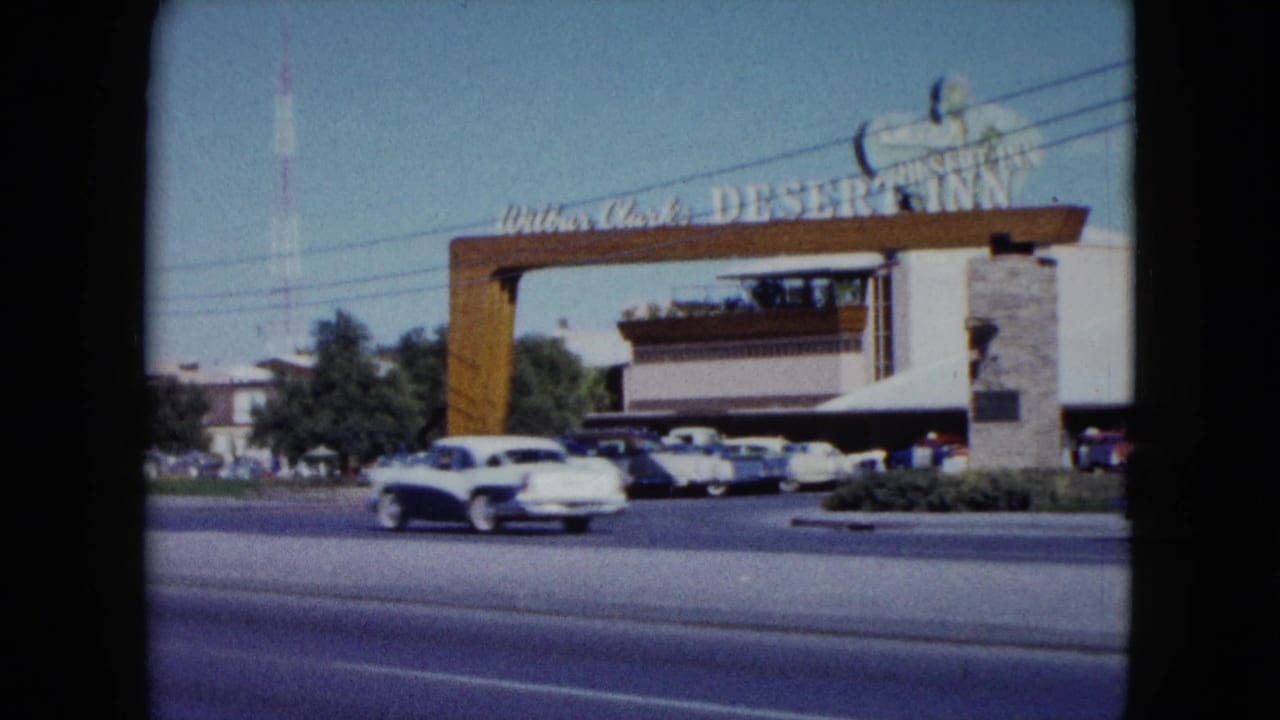 A white mid-century car passes the Desert Inn entrance in Nevada, with the arch, parking lot, building, and roadside landscaping visible behind it.