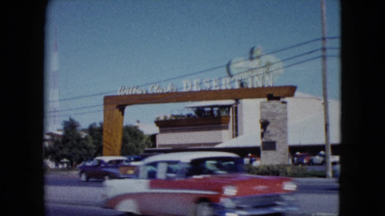 A red and white mid-century car crosses the foreground in motion blur while the Desert Inn entrance, roadside lanes, parked cars, and tower remain visible behind it.