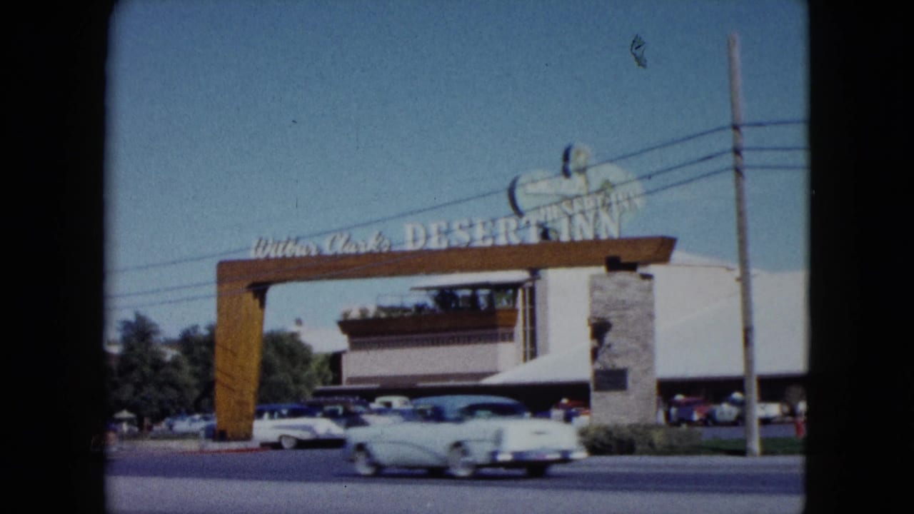1959 Nevada 8mm frame showing the Desert Inn entrance sign, stone pillars, parked cars, and a white sedan crossing the foreground road.