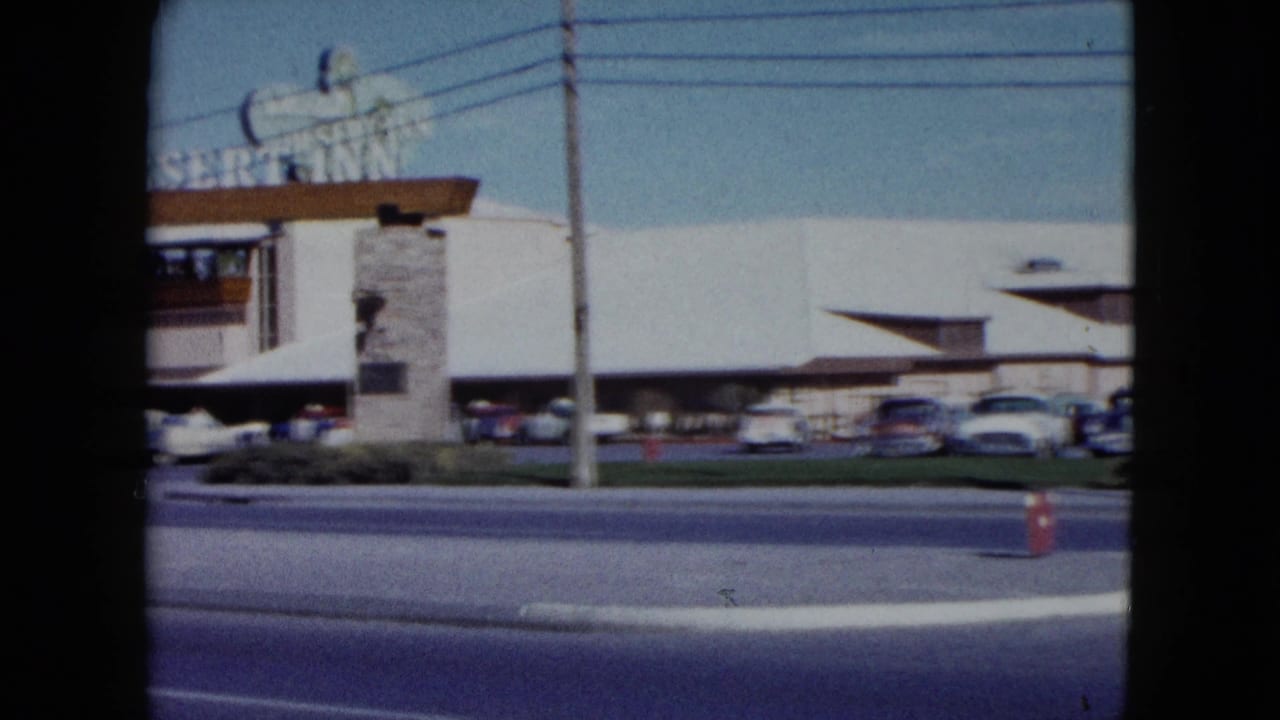 1959 Nevada frame with part of the Desert Inn sign at left, the resort facade, parked cars, roadway, utility pole, and an orange cone.