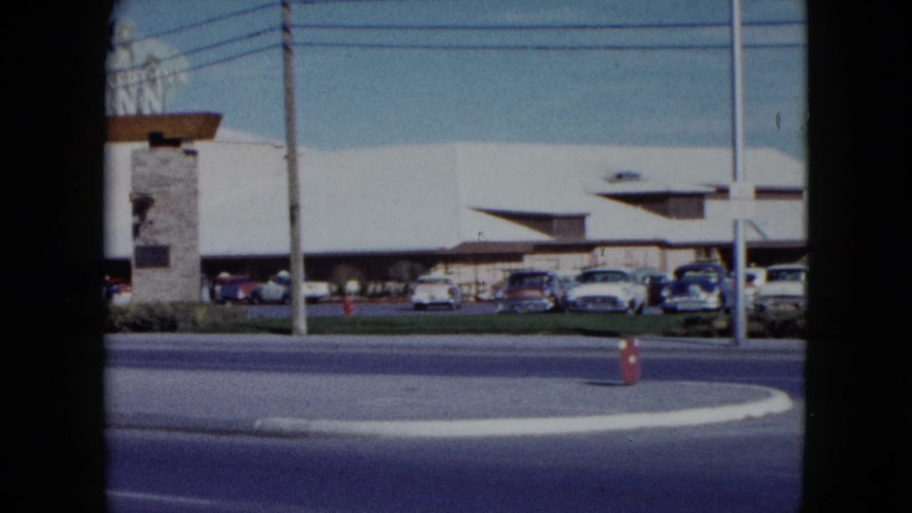 1959 Nevada roadside view with parked cars, moving traffic, utility lines, and a low mid-century building across the street.