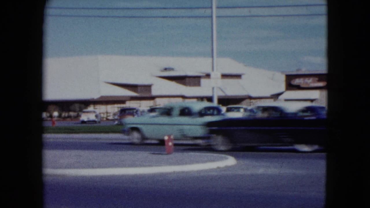 A light blue car and dark car pass in front of a parking area and roadside building.