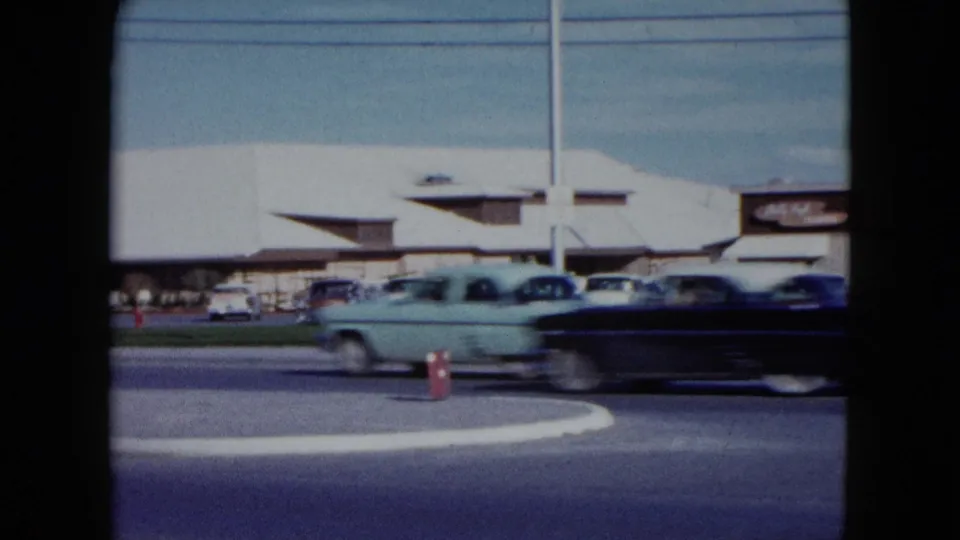 Moving 1950s cars cross the frame in front of a Nevada roadside parking area and low modern building.