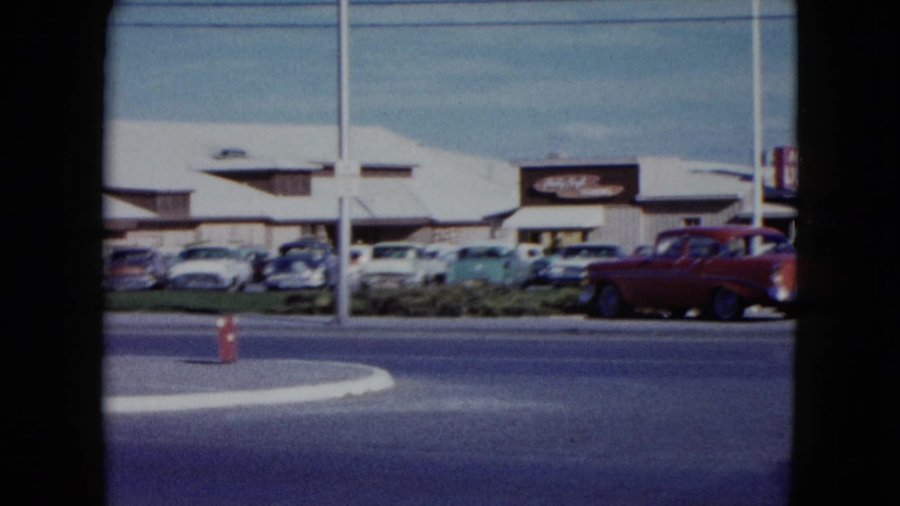 1959 Nevada roadside parking lot with rows of cars, a red car in motion, utility poles, and visible commercial signage.