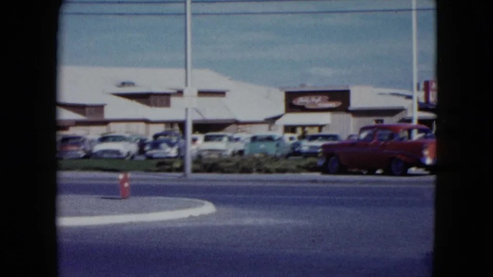 1959 Nevada roadside parking lot with rows of cars, a red car in motion, utility poles, and visible commercial signage.