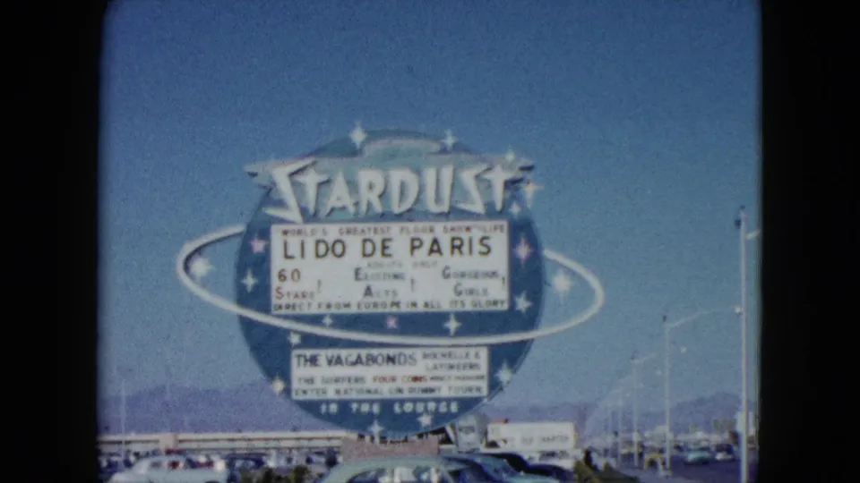 Large Stardust Hotel sign advertising Lido de Paris in Nevada, with cars, roadside poles, blue sky, and distant mountains.