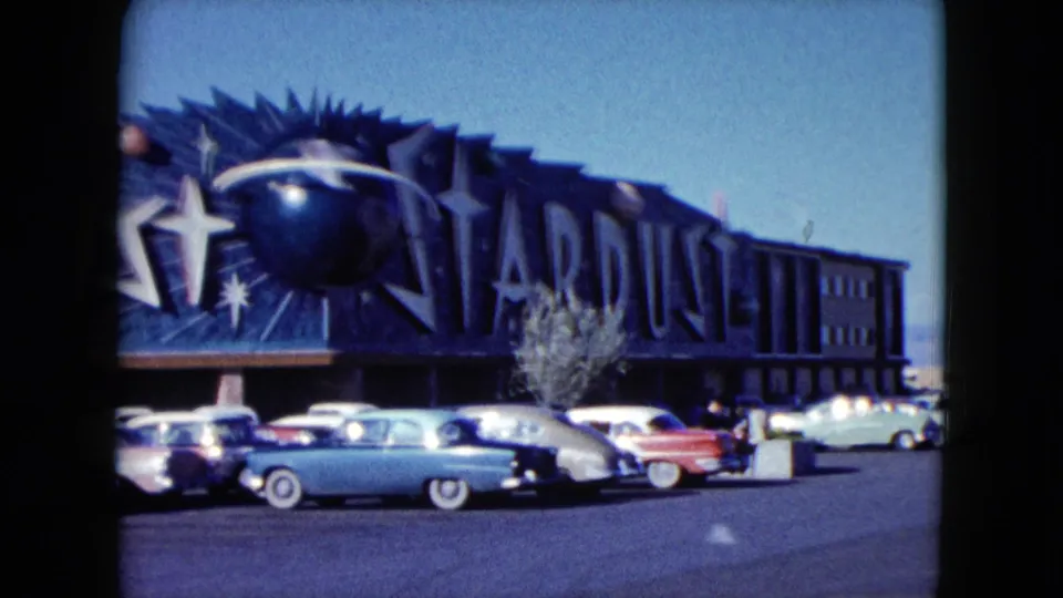 1959 Nevada 8mm frame showing a closer view of the Stardust sign, star graphics, pastel cars, and sunlit parking lot.