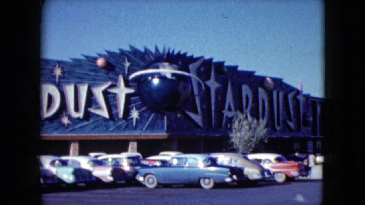 1959 Nevada frame showing the Stardust sign across the building, starburst graphics, a row of cars, and a small tree in front.