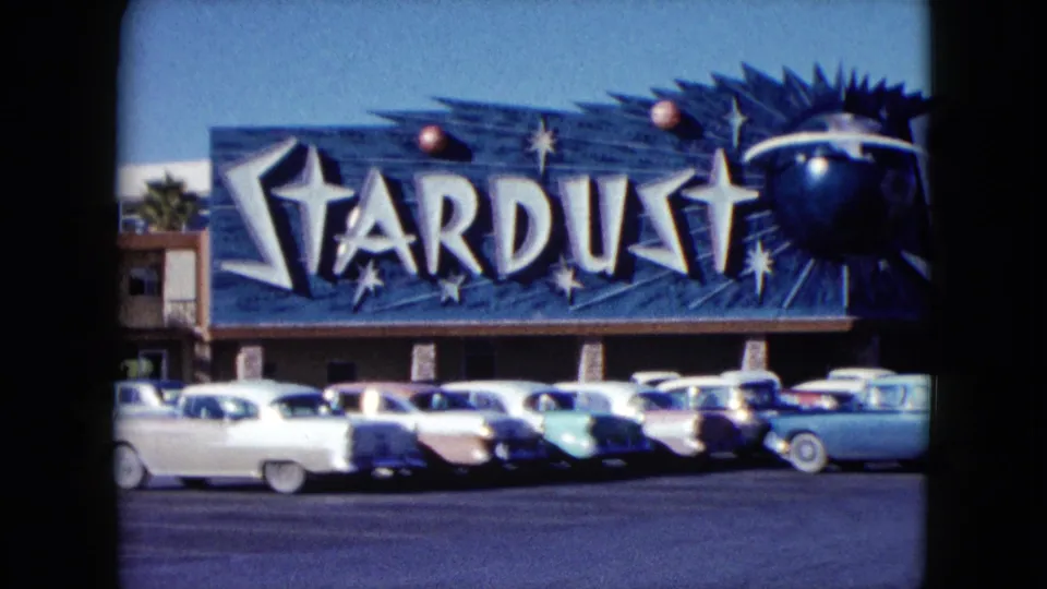 1959 Nevada frame with the Stardust sign partly cropped, low motel buildings, palm tree, and parked late-1950s cars.