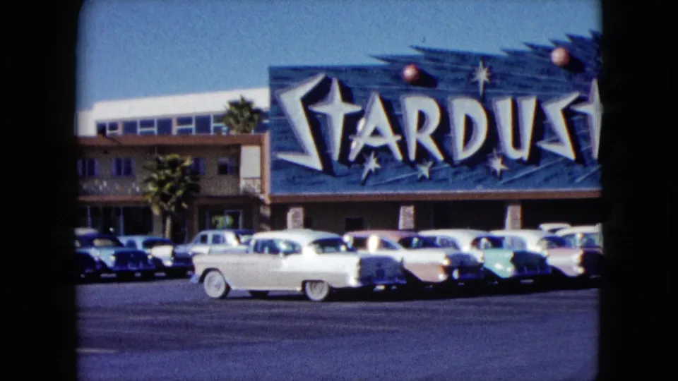 A white classic car crosses the foreground before the Stardust sign and parked vehicles in a 1959 Nevada home movie frame.