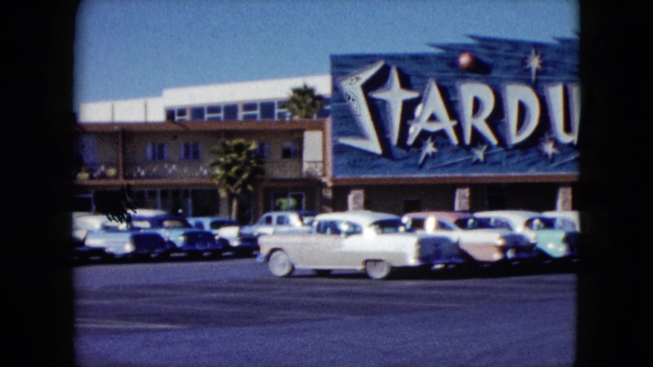 1959 Nevada home-movie frame showing the Stardust casino sign, parked cars, palm trees, and a white car crossing the lot.