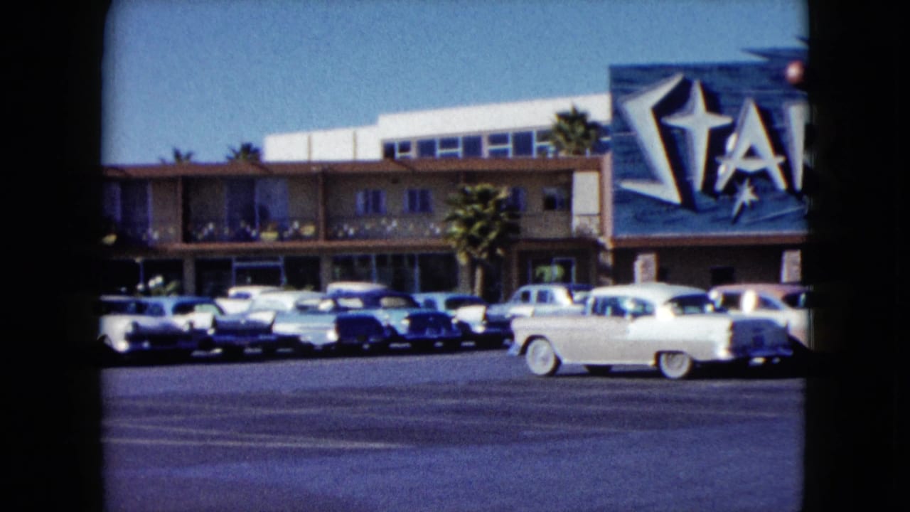 White car drives past rows of parked cars in front of the Stardust sign.