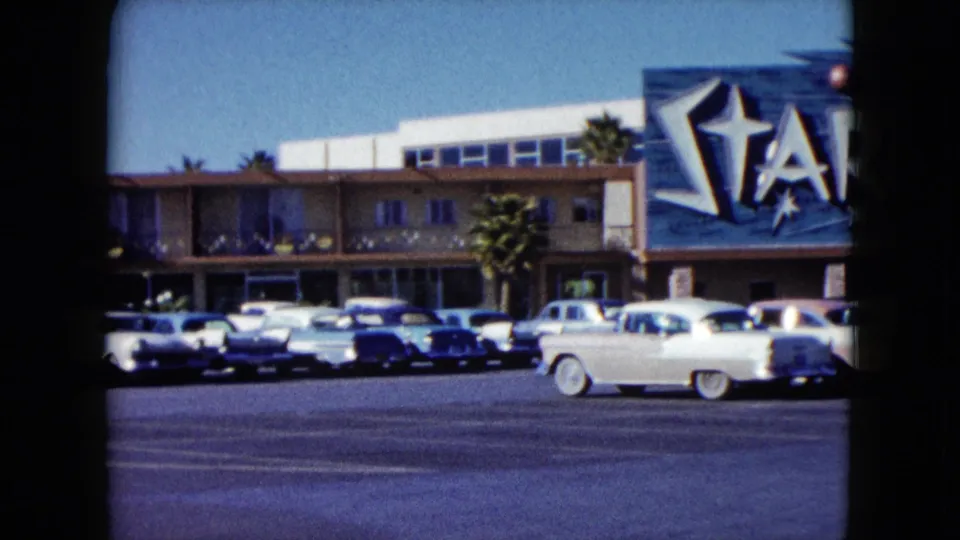 1959 Nevada frame with a white car moving through the Stardust casino parking lot below the large sign and building facade.