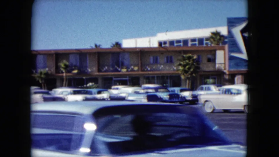 1959 Nevada home-movie frame with a blurred passing car in front of parked vehicles and the Stardust building edge.