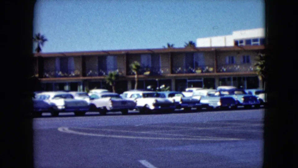1959 Nevada frame showing the Stardust casino frontage, parked late-1950s cars, palm trees, and open asphalt under a clear sky.