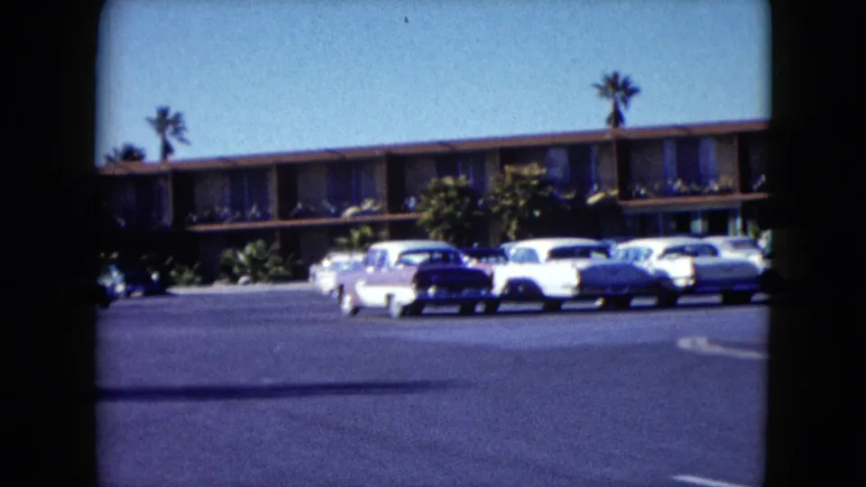 1959 Nevada travel footage frame with two-tone cars, motel frontage, palms, and open asphalt lot.
