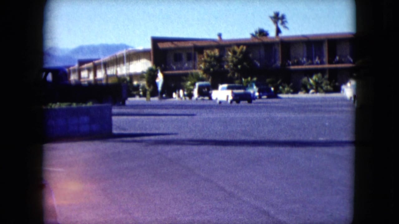 Motel building with parked cars, palm trees, and distant hills.