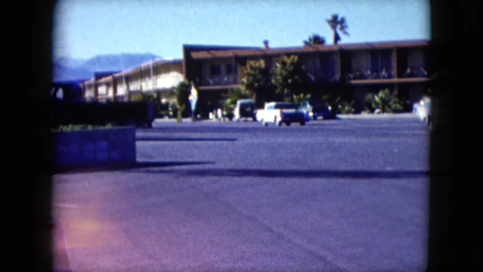 1959 Nevada motel parking view with two-tone cars, palm landscaping, distant hills, and small human figures near the building.
