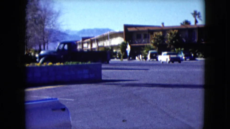 1959 Nevada 8mm frame with a dark truck in the foreground, motel rooms, classic cars, a planter, and distant people in the lot.