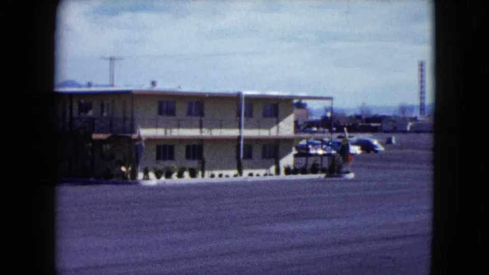 1959 Nevada frame showing another low-rise motel building, exterior stairs, parked cars, open asphalt, and distant urban structures.