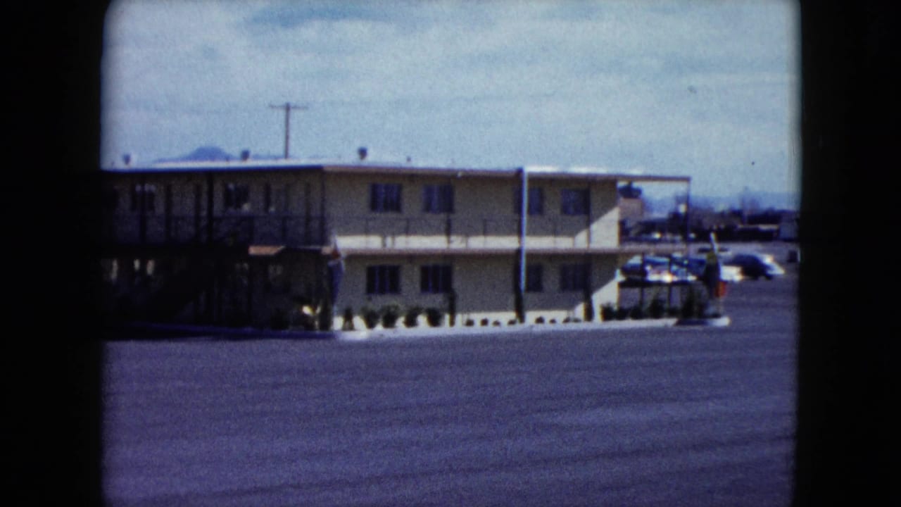 1959 Nevada home-movie frame showing a two-story roadside motel across a broad paved parking lot, with utility poles and hills beyond.