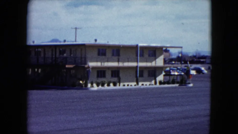 1959 Nevada home-movie frame showing a two-story roadside motel across a broad paved parking lot, with utility poles and hills beyond.