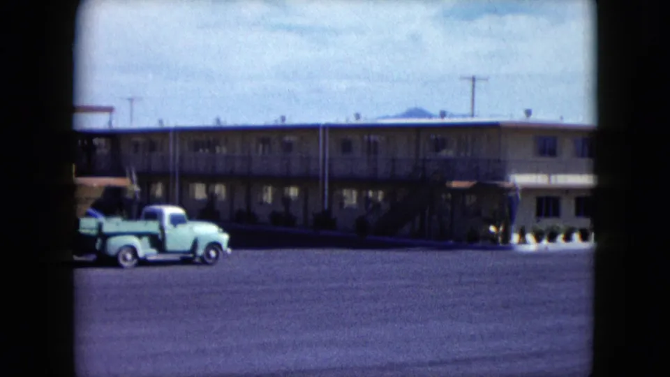 1959 Nevada home-movie frame with a light green vintage pickup truck moving across the motel parking lot in front of a two-story building.