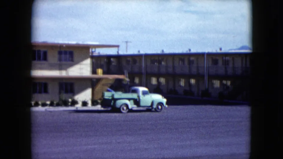 1959 Nevada frame showing a light green pickup truck near the motel frontage, with balcony walkways, stairs, windows, and distant hills visible.