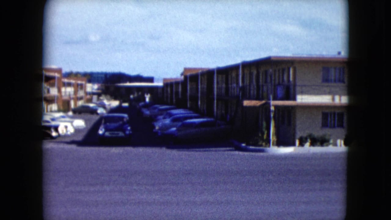 1959 Nevada 8mm frame showing a motel parking lot, exterior corridors, parked mid-century cars, and a blue pickup in afternoon light.