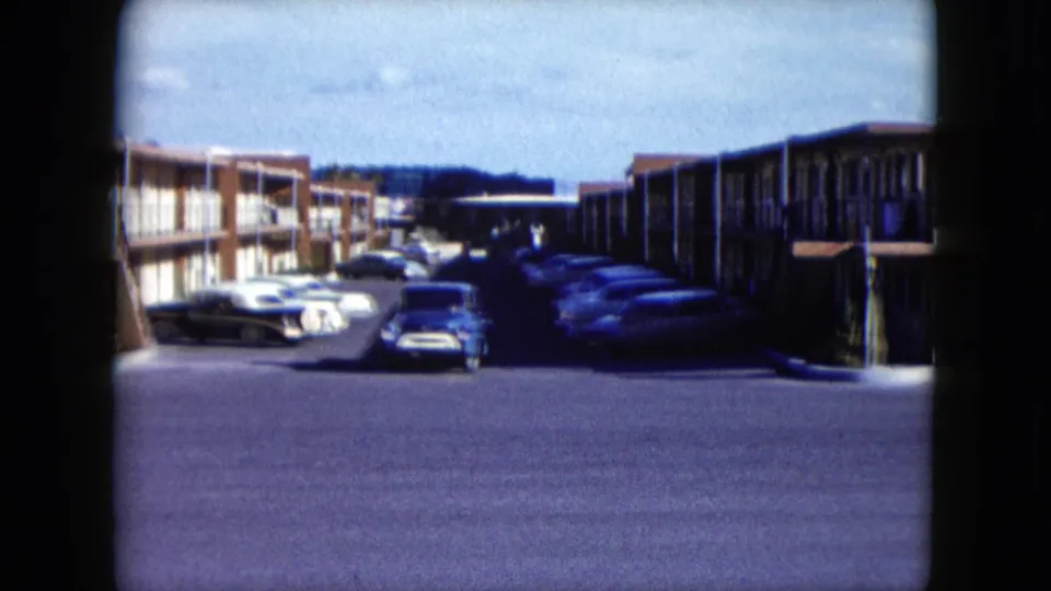 1959 Nevada motel lot seen on 8mm film, with exterior room rows, several parked cars, and a blue pickup facing the camera.