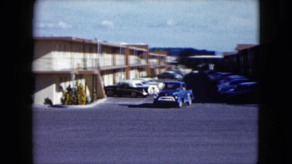 1959 Nevada home-movie frame with a blue pickup crossing a motel lot beside parked cars and exterior motel balconies.