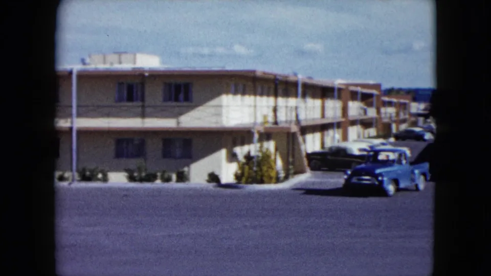 1959 Nevada 8mm frame showing the blue pickup near the right side of a motel parking lot, with rooms, stairs, shrubs, and parked cars behind it.