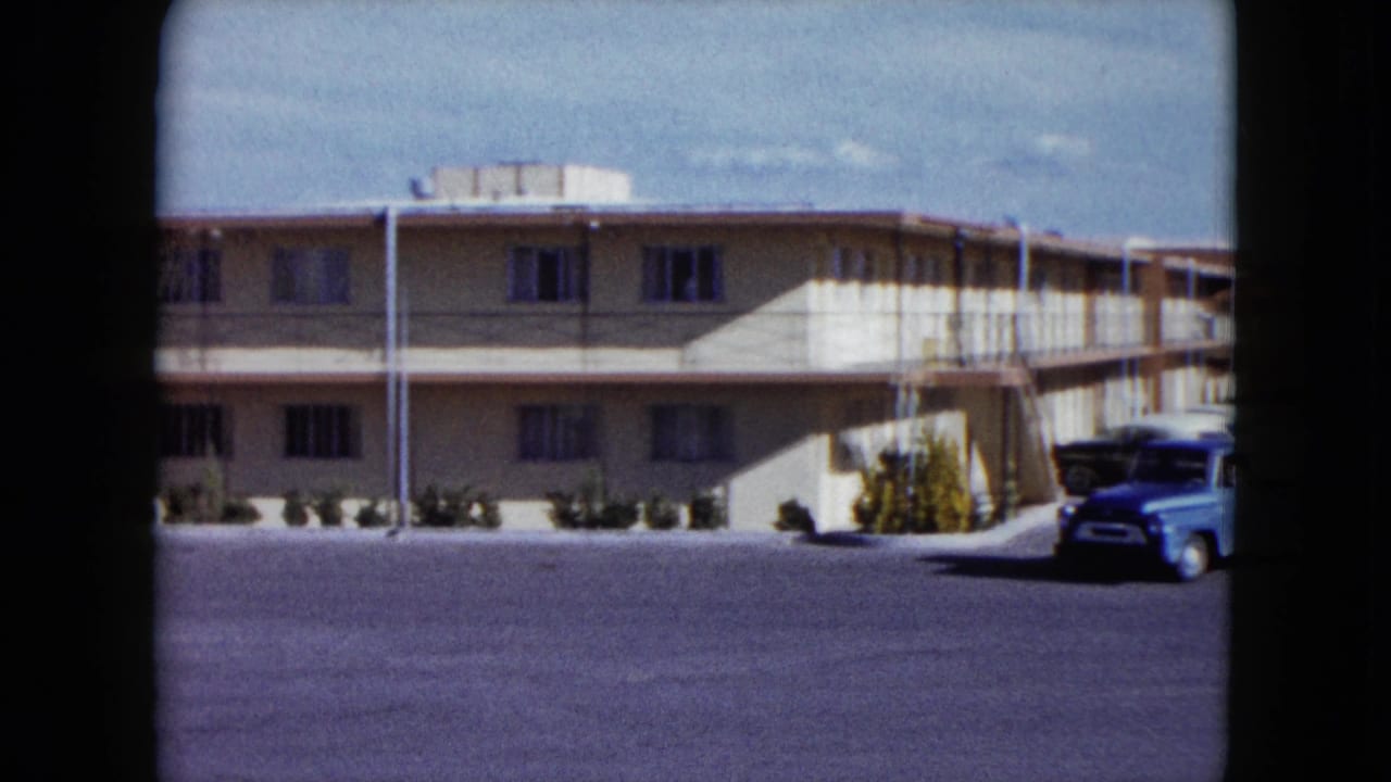 1959 Nevada home-movie frame showing a beige roadside motel, balcony walkway, shrubs, and a light blue pickup partly entering at right.