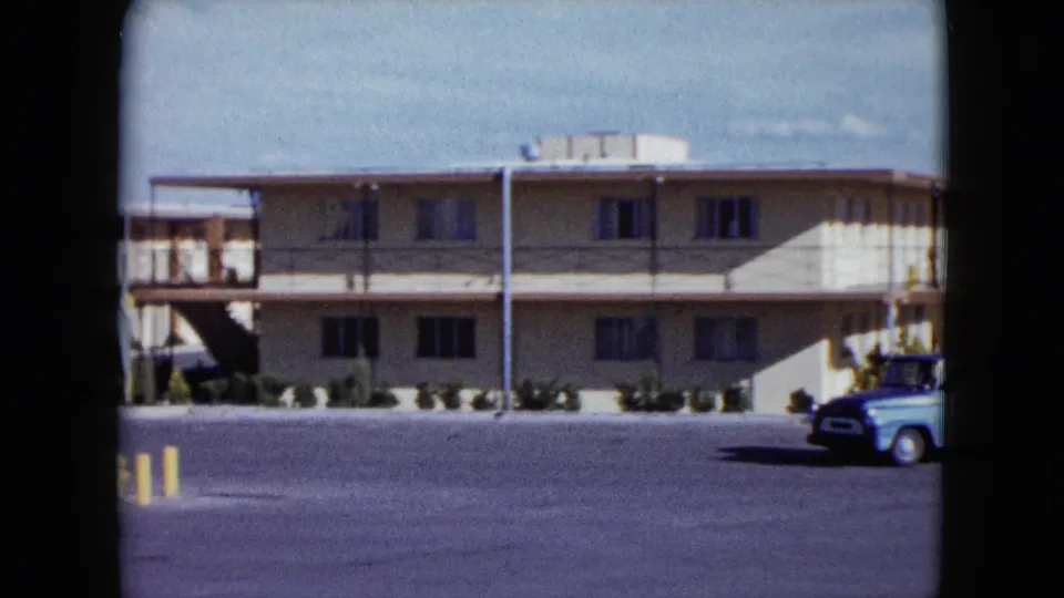 1959 Nevada frame of a roadside motel exterior with balcony railings, yellow parking-lot bollards, and a light blue pickup near the right edge.
