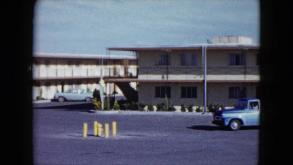 1959 Nevada home-movie still with a light blue pickup crossing the foreground of a beige two-story motel and open parking lot.
