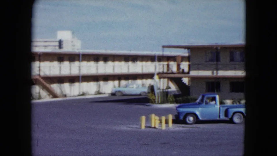 1959 Nevada frame showing a light blue pickup in the motel parking lot, with a beige two-story roadside motel and balcony behind it.