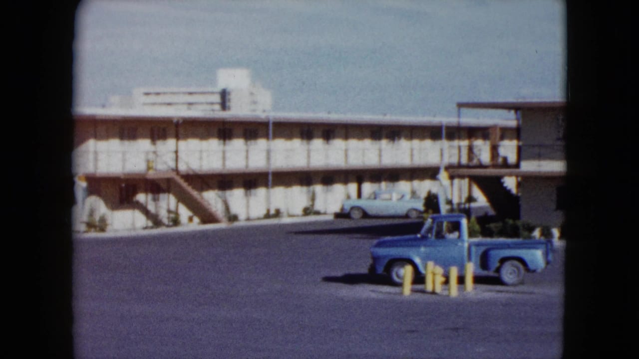 1959 Nevada home-movie frame showing a blue pickup in a motel parking lot with exterior stairs and a pale sedan nearby.