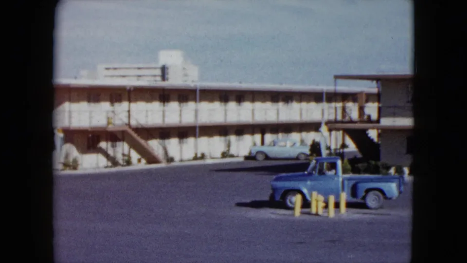 1959 Nevada home-movie frame showing a blue pickup in a motel parking lot with exterior stairs and a pale sedan nearby.