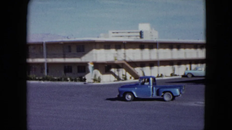 Blue pickup seen from a static 8mm view in a Nevada motel lot, with exterior stairs and a parked light sedan in the background.