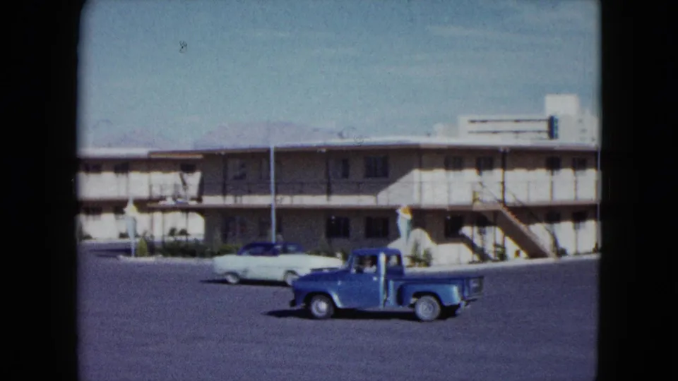 1959 Nevada roadside motel view with a blue pickup passing a pale sedan in a broad parking area.