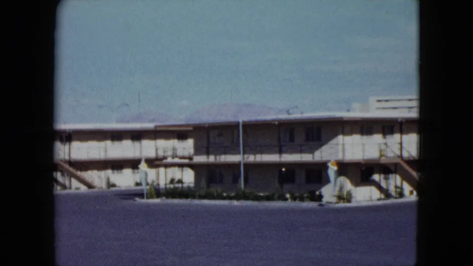 Static 8mm frame of a Nevada motel frontage with exterior stairs, planted strip, open asphalt, and distant figures or details near the building.