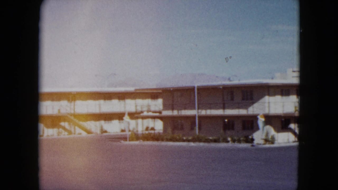 1959 Nevada home-movie frame showing a long motel facade across an open parking lot with exterior stairs and walkway.